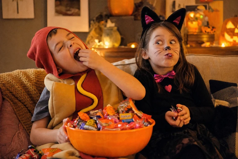 A photo of two kids in costumes eating Halloween candy. The scene should be silly, fun, and warm