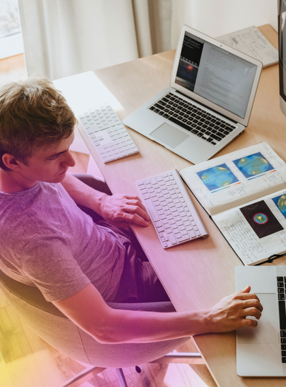 Man Working Young man seated at a desk working on two laptops and notebooks with diagrams and keyboards nearby.