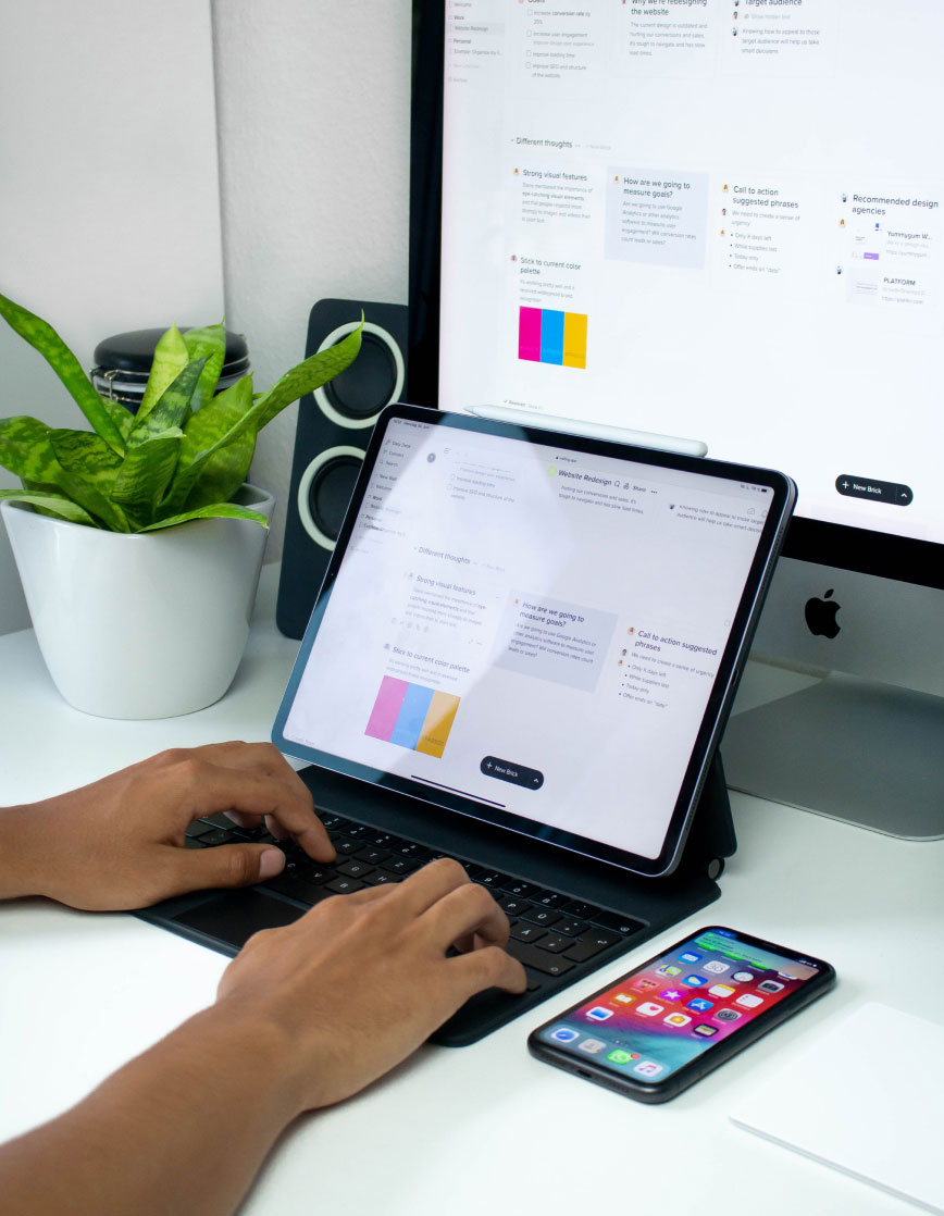 Person Working on Tablet Person typing on a tablet with keyboard on a desk, iPhone beside it, monitor and potted plant in the background.
