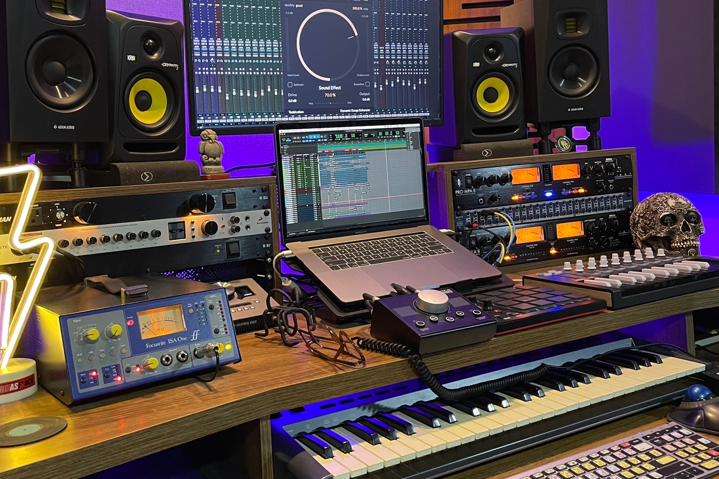 Home music studio desk with laptop running a DAW, studio monitors, MIDI keyboard, audio interfaces, controllers, rack gear and a decorative skull.