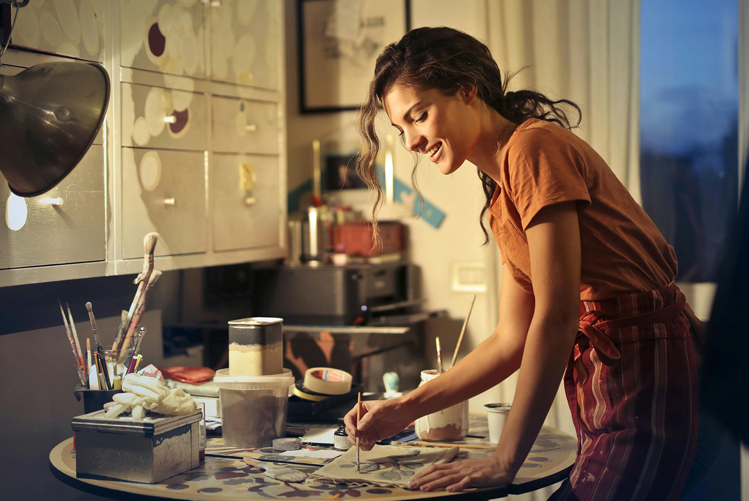 woman-painting-in-a-art-studio Young woman in an apron smiling while painting at a cluttered, lamp-lit craft table with brushes and jars.